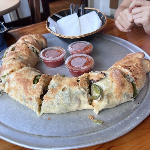 A large, crescent-shaped calzone, sliced and served with three small cups of marinara sauce on a metal tray.