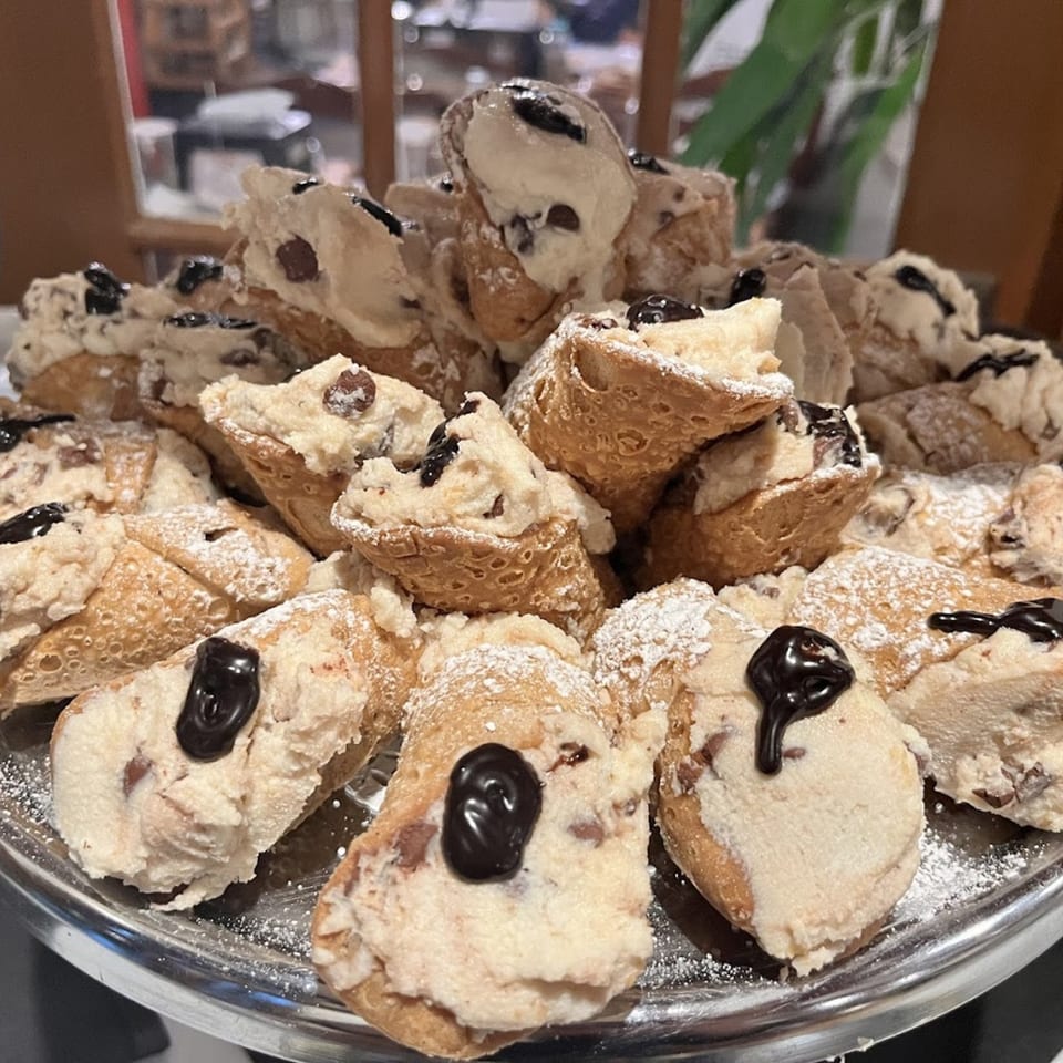 A silver platter piled high with cannolis, dusted with powdered sugar and drizzled with chocolate.