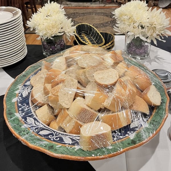 Sliced bread on a decorative platter, covered in plastic wrap, with flowers and plates in the background.